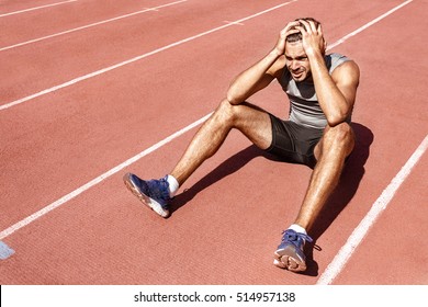 Dealing With The Loss. Young Male Athlete Looking Upset And Disappointed After Losing The Race Sitting On The Racetrack With His Hands To His Head Copyspace