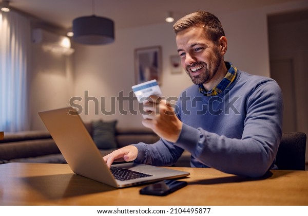 Debit, credit card,\
online banking, and shopping. A happy young man is sitting in his\
living room at home, holding a credit card and typing a card number\
on the laptop.