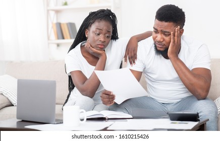 Debt Concept. Portrait Of Sad Afro Couple Looking At Invoice, Upset About Huge Bills, Not Able To Pay Mortgage, Sitting Depressed On Sofa At Home