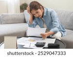 Debt and financial concept, stressed asian young woman have a headache while female sitting on sofa calculate expenses, income, hand holding bills for prepare payment or pay bills on table at home.