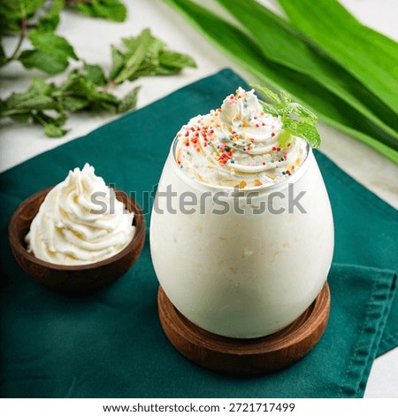 A decadent vanilla milkshake served in a rounded glass, topped with swirls of whipped cream, colorful sprinkles, and a fresh mint leaf. A small wooden bowl of extra whipped cream is placed beside it, perfect for a sweet treat.
