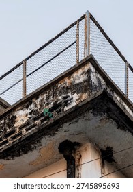 Decayed concrete building corner with rusted fence, water damage, mold stains, and peeling paint, illustrating urban decay and neglected architecture in industrial settings.