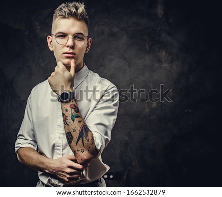 Decisive young student guy, with tattoos on his hand, wearing white shirt, glasses and watches, posing in a dark studio isolated on grey background, looking confident and thoughtful
