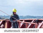 A deck crew member prepares the forward mooring station for docking, focused on securing the ship safely as it approaches the port. The intensity of the task contrasts with the calm surroundings.