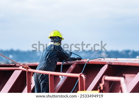 A deck crew member prepares the forward mooring station for docking, focused on securing the ship safely as it approaches the port. The intensity of the task contrasts with the calm surroundings.
