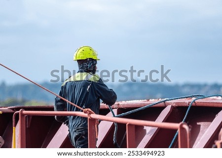 A deck crew member prepares the forward mooring station for docking, focused on securing the ship safely as it approaches the port. The intensity of the task contrasts with the calm surroundings.