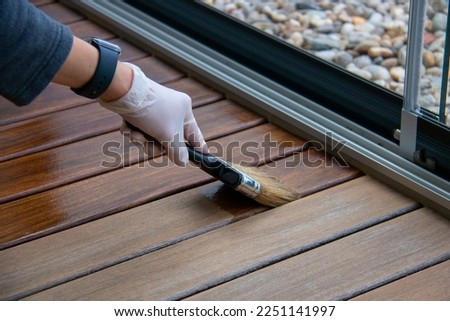 Deck staining, worker applying deck oil on decking boards with paint brush, hardwood terrace renovation