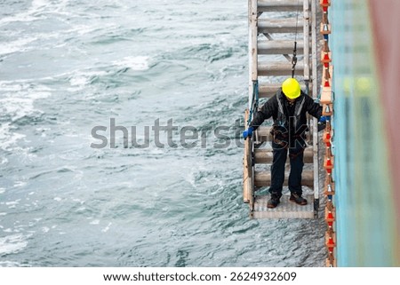 A deckhand prepares the pilot ladder as the ship readies for port entry. Secured in a safety harness, he carefully ensures the setup is ready, focusing on the task in a high-risk environment.