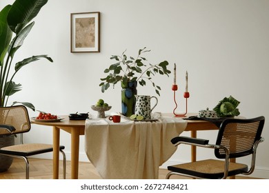 Decorated dining table with eucalyptus, patterned ceramics, red candlesticks, limes, tomatoes, and rattan chairs in modern interior.