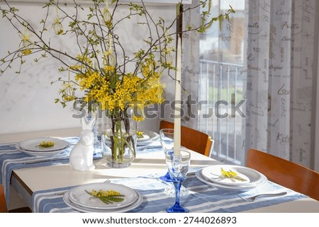 Decorated dining table prepared for an easter celebration. Featuring a centerpiece with mimosa branches. Decorative easter egg ornaments. A ceramic bunny. And place settings with blue striped runners
