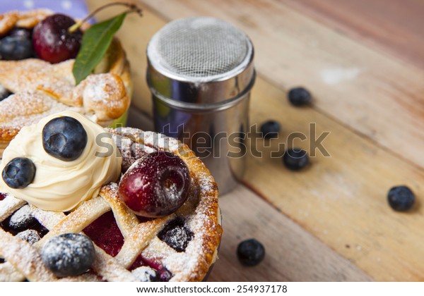Decorated homemade shortcrust pastry berry\
pie with polka dot cloth, shiny metal icing sugar shaker and\
selection of berries on grunge style wooden\
table