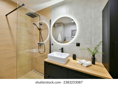 El cuarto de baño es moderno y está decorado al estilo boho, con una cabina de ducha a ras de suelo, una columna de ducha de efecto lluvia y armarios negros empotrados, con baldosas imitación de madera color miel y un espejo redondo de efecto luminoso.