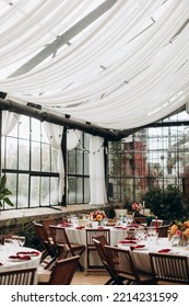 A Decorated Party Hall With Many Windows And White Capes. Numbered And Decorated Tables With Transparent Glasses, White Dishes And Red Napkins.	