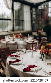 Decorated Served Table With A Decorative Bouquet Of Flowers In The Middle. A Round Table With A White Tablecloth And White Dishes With A Red Napkin Tied In A Bow, Transparent Glasses And Silver Cutler