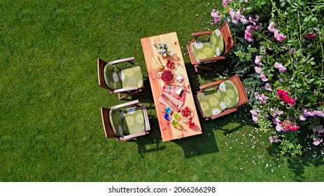 Decorated table with cheese, strawberry and fruits in beautiful summer rose garden, aerial top view of table food and drinks setting outdoors from above. Leisure and picnic with family and friends