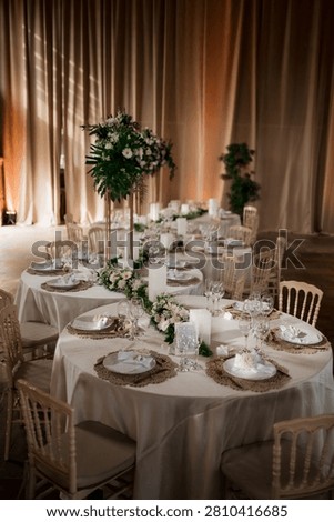 A decorated wedding reception table featuring white linen, tall floral centerpieces with lush greenery, pillar candles, and stylish place settings.