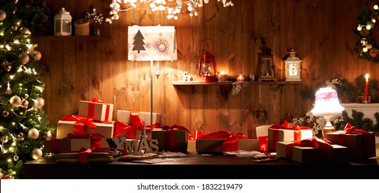 Decorated xmas table with Merry Christmas gifts in cozy Santa home interior, banner. Happy New Year presents boxes in workshop late in night with lights on xmas tree, holiday eve background.