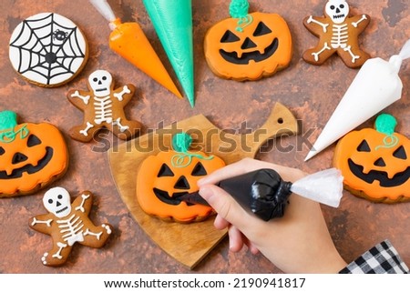 Decorating Halloween gingerbreads of pumpkin lantern and skeletons with frosting. Girl holds pastry bag with black icing and decorates gingerbread pumpkin lantern