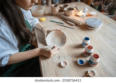 Decorating pottery. Woman ceramist holding paintbrush and drawing cute ornament on clay plate. Creative classes of work with earthenware, creating an authors style in tableware design. Artist at work. - Powered by Shutterstock