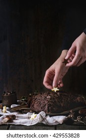 Decorating process of homemade Christmas chocolate yule log by woman's hands with chocolate stars over old wooden table with holly branch and chestnuts. Dark rustic style.