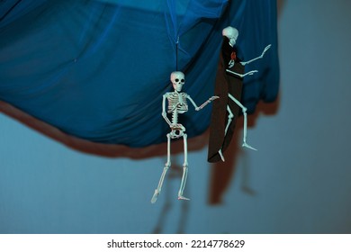 Decoration For Halloween: Two Skeletons Hanging From A Blue Cloth On A Blue Background In A Dark Room. One Skeleton In A Black Cape With A Red Bow-tie.