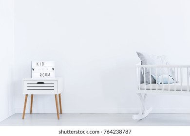 Decoration On White Cabinet Next To White Cradle In Empty Interior With White Wall