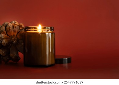 A decorative aromatic candle in a glass jar, accompanied by a natural pinecone, all set against a bold red background - Powered by Shutterstock