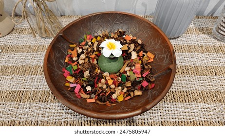 Decorative centerpiece with a white frangipani flower on a green ceramic base surrounded by colorful dried potpourri in a brown leaf etched bowl on a woven mat, creating a cozy and inviting atmosphere - Powered by Shutterstock