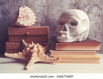 A decorative figure in the form of a human skull is on a stack of books. Nearby is a wooden box on top of which is a seashell. In the foreground is a starfish. Stone or cement background with cracks. - Powered by Shutterstock