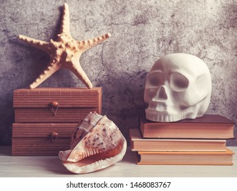 A decorative figure in the form of a human skull is on a stack of books. Nearby is a wooden box on top of which is a starfish. In the foreground is a seashell. Stone or cement background with cracks. - Powered by Shutterstock