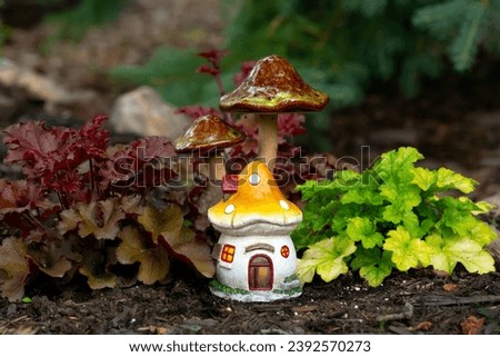 Decorative garden miniature ceramic figurine of a fairy house and mushrooms are among red and green heuchera plants in the flower bed.