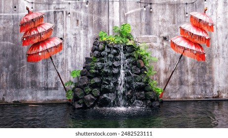 A decorative garden waterfall surrounded by traditional balinese red umbrellas, creating a tranquil and cultural outdoor ambiance - Powered by Shutterstock