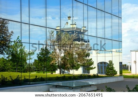  Decorative mirror facade of the building with a reflection of the neighboring building, park area and cloudy sky in it. Copy space.                                         