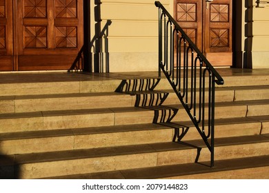 decorative painted black steel stair railing. open exterior space. wide beige stone stairs. elegant staines oak entrance doors with ornate carved pattern. bright sunlight. strong shadows. perspective.
