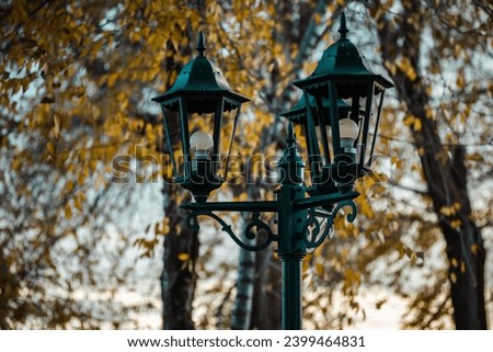 Decorative retro cast-iron lantern with three lamps in autumnal park. Antique lamp post outdoors in autumn. Beautiful street lamp in fall season against tree with yellow leaves Dark key nature photo 