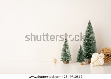 Decorative side view setup with mini snow-covered Christmas trees and paper ornaments in a Scandinavian minimalist style, placed against a neutral backdrop