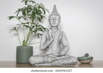 Decorative statuette of Buddha in meditative position on a wooden table, next to a pot with plants and a small bowl containing a green stone. White background - Powered by Shutterstock