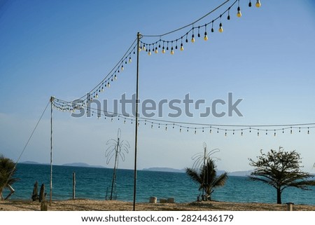 Decorative string lights are strung across a sandy beach, overlooking the calm turquoise ocean under a bright blue sky