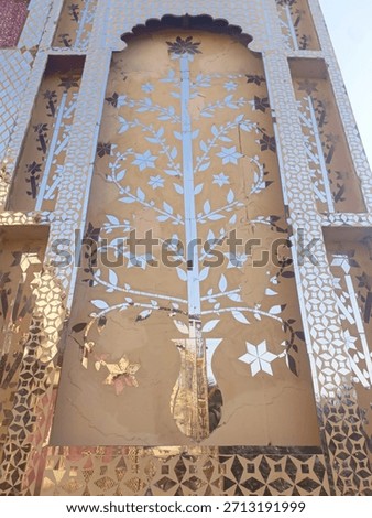 A decorative wall panel featuring intricate mirror work, floral patterns, and a tall tree design, showcasing traditional craftsmanship and artistic architectural detailing in warm lighting.
