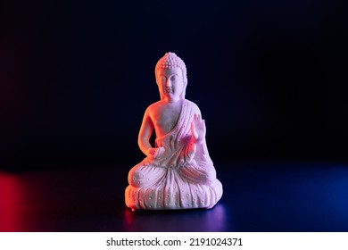 Decorative White Buddha Statuette On The Dark Background In Red And Blue Neon Light. Duality Concept, Yin Yang. Internal Struggle Of Opposites. Selective Focus, Copy Space.