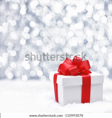 Decorative white gift box with a large red bow standing in fresh snow against a background bokeh of twinkling party lights