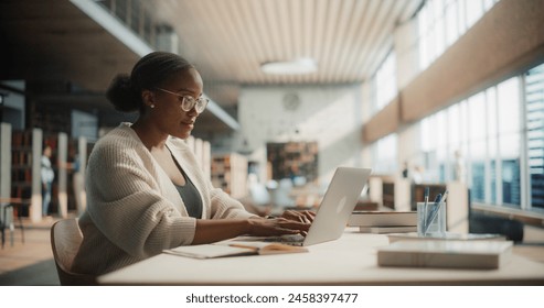 Dedicated African American Female Student Engaged in Online Learning at a Modern Library. Young Woman Using Laptop for Research, Surrounded by Books and Academic Environment. - Powered by Shutterstock