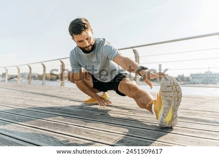 A dedicated athlete stretches on a boardwalk, focusing intently on his form against a backdrop of a gentle river and city skyline.

