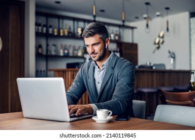 A dedicated busy man dressed in smart casual is sitting in working friendly cafe and typing a report on the laptop. He has earphones in his ears. A businessman typing on a laptop in a cafe.