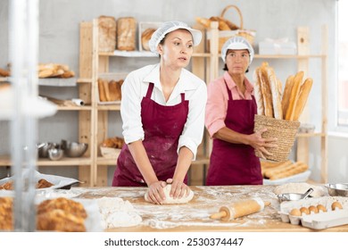 Dedicated female baker in maroon apron kneading pliable dough on flour-covered work surface in cozy artisan bakery against backdrop of wooden shelves lined with bread loaves, baguettes and pastries - Powered by Shutterstock