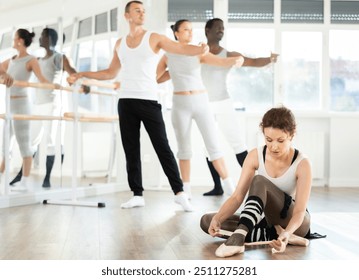 Dedicated female dancer sitting on floor in spacious sunlit choreography studio and adjusting point shoes, preparing for ballet class with dancers practicing at barre in background.. - Powered by Shutterstock