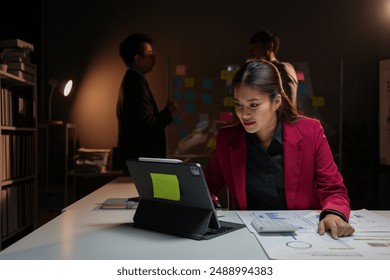 Dedicated female professional working late in dimly lit office using digital tablet while colleagues hold a meeting in the background - Powered by Shutterstock