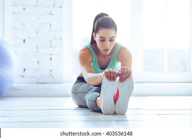 Dedicated To Fitness. Front View Of Beautiful Young Woman In Sportswear Doing Stretching While Sitting On The Floor In Front Of Window At Gym