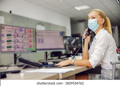 Dedicated Hardworking Blond Female Boss In Suit With Face Mask On Sitting In Control Room In Heating Plant And Having Important Phone Conversation During Corona Virus Outbreak. Womam At Work.