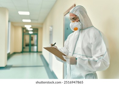 A dedicated healthcare worker, fully equipped with protective gear, is diligently reviewing important patient information while standing in a busy hospital corridor during these challenging times - Powered by Shutterstock